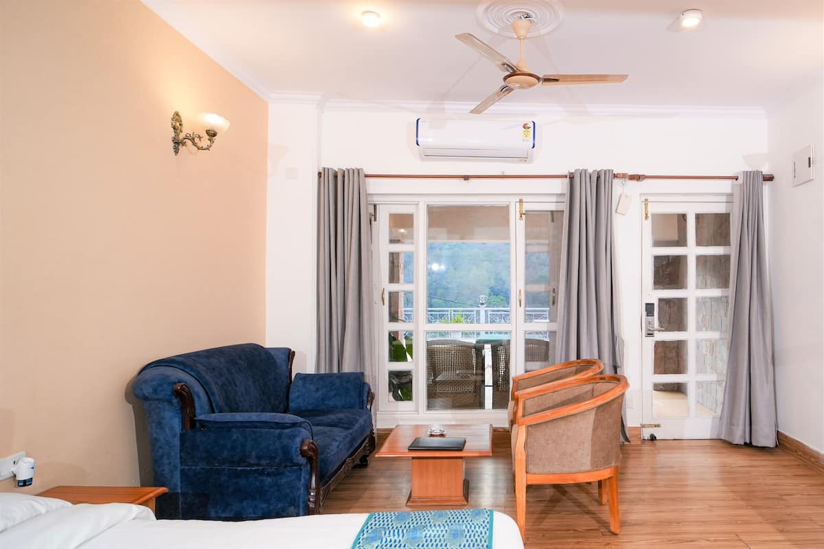 A hotel room with a bed, a blue armchair, a wooden chair, a small table, and a ceiling fan.