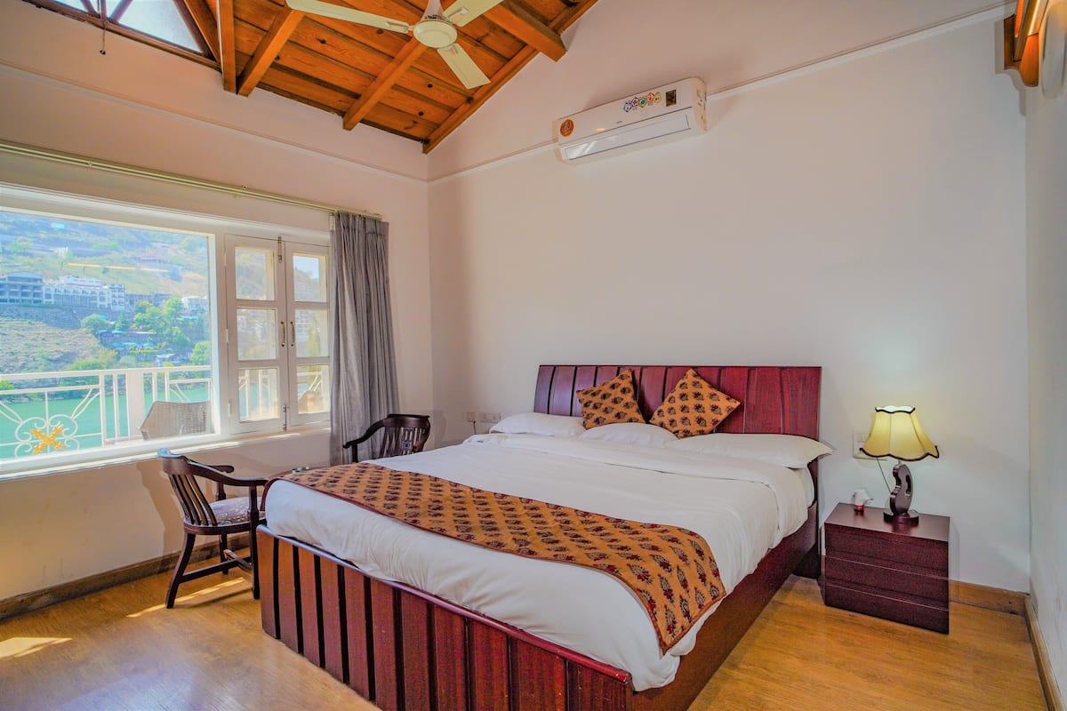 A bedroom with a bed, a chair, a nightstand, a lamp, and a window with a view.