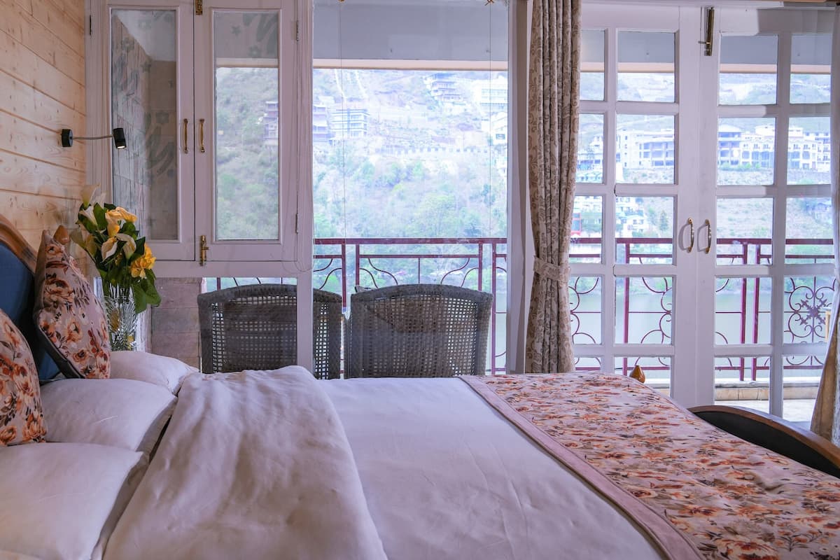 A bedroom with a bed, floral pillows, a vase with flowers, wicker chairs, and a balcony with a railing.