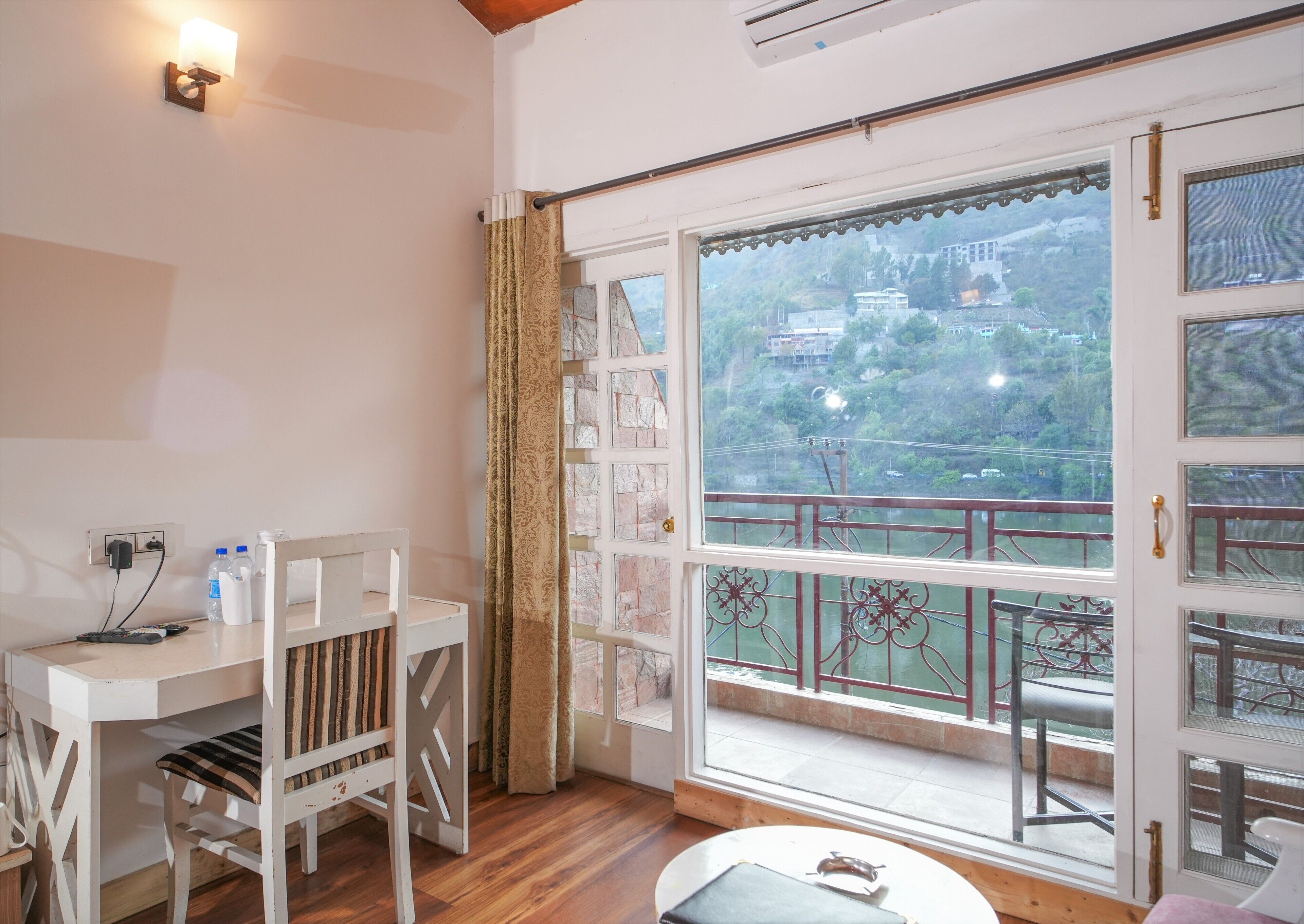 Una habitación con piso de madera, una mesa blanca con una silla, un ventanal con vistas a un paisaje montañoso y un balcón con barandilla.