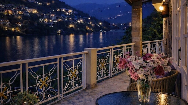 A terrace with a table and chairs, a railing, and a view of a lit-up town across a body of water. - Neelesh Inn - Luxury Lake View Hotel With Swimming Pool On Bhimtal Lake (Nainital)