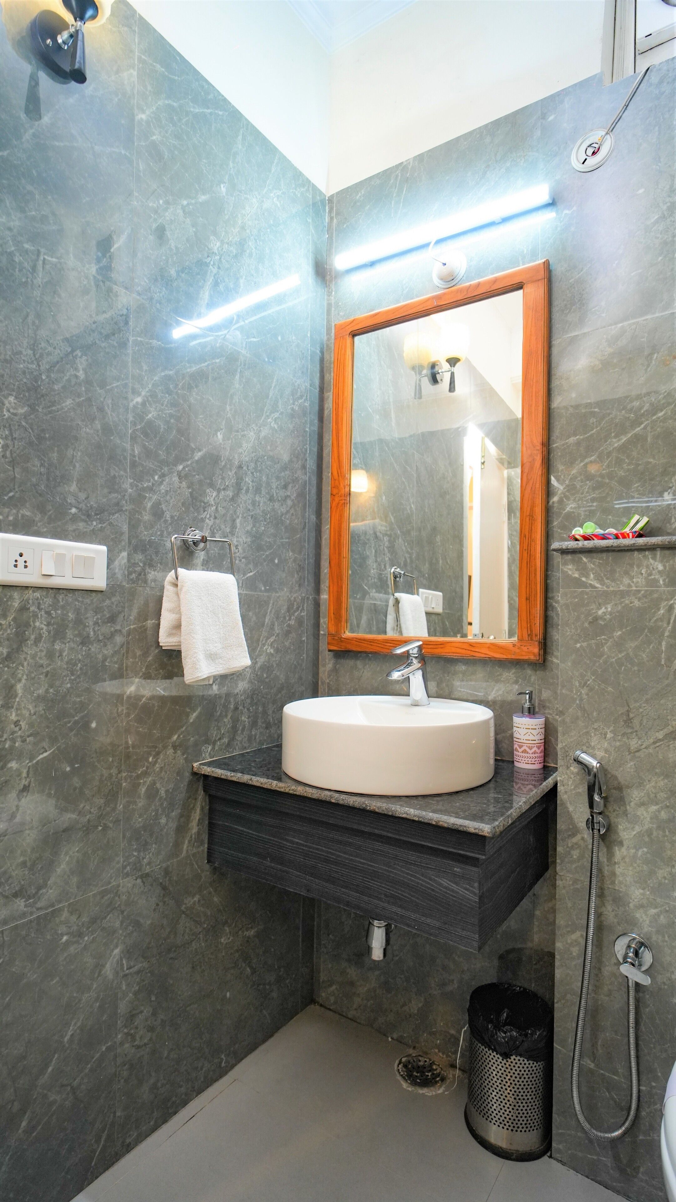 Un baño con pared de mármol, espejo con marco de madera, lavamanos blanco, cabezal de ducha de mano y toallero.