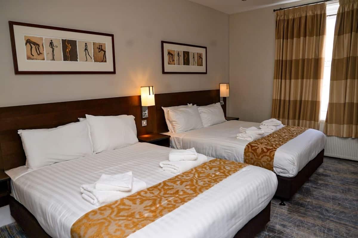 Une chambre d’hôtel avec deux lits, chacun agrémenté de draps blancs et d’un couvre-lit à motifs. Les murs sont ornés d’œuvres d’art encadrées, il y a une fenêtre avec des rideaux et une table de chevet avec une lampe.