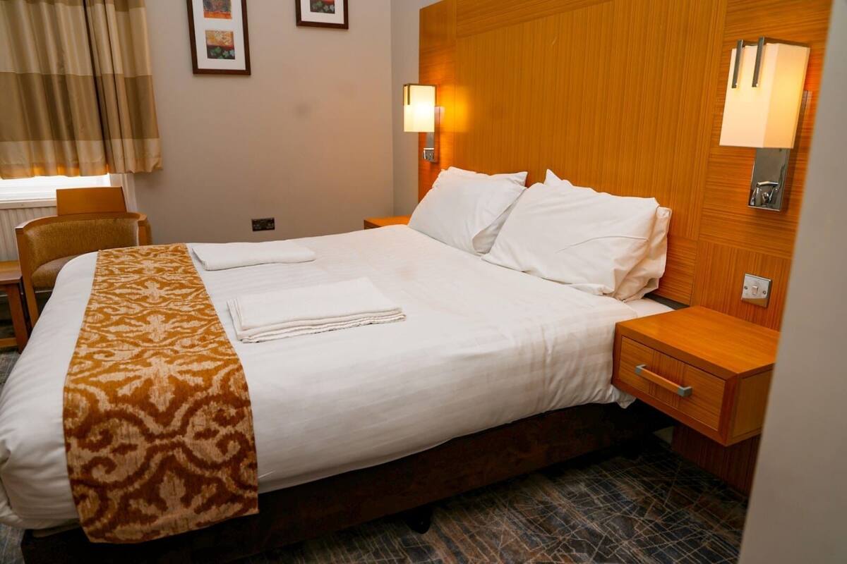 Une chambre d’hôtel avec un grand lit, une tête de lit en bois, deux tables de chevet et un plaid à motifs.
