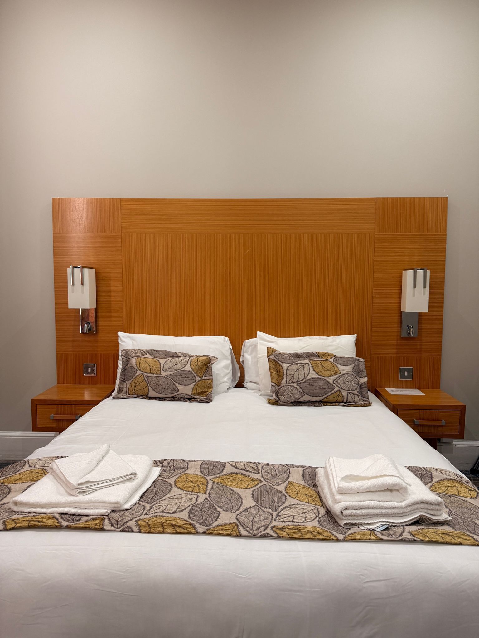 A neatly made bed with a patterned bedspread, two pillows, and folded towels on a wooden headboard with wall-mounted lamps.