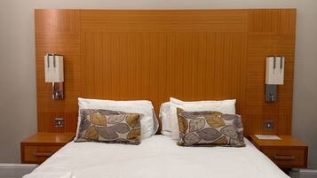 A neatly made bed with a patterned bedspread, two pillows, and folded towels on a wooden headboard with wall-mounted lamps.