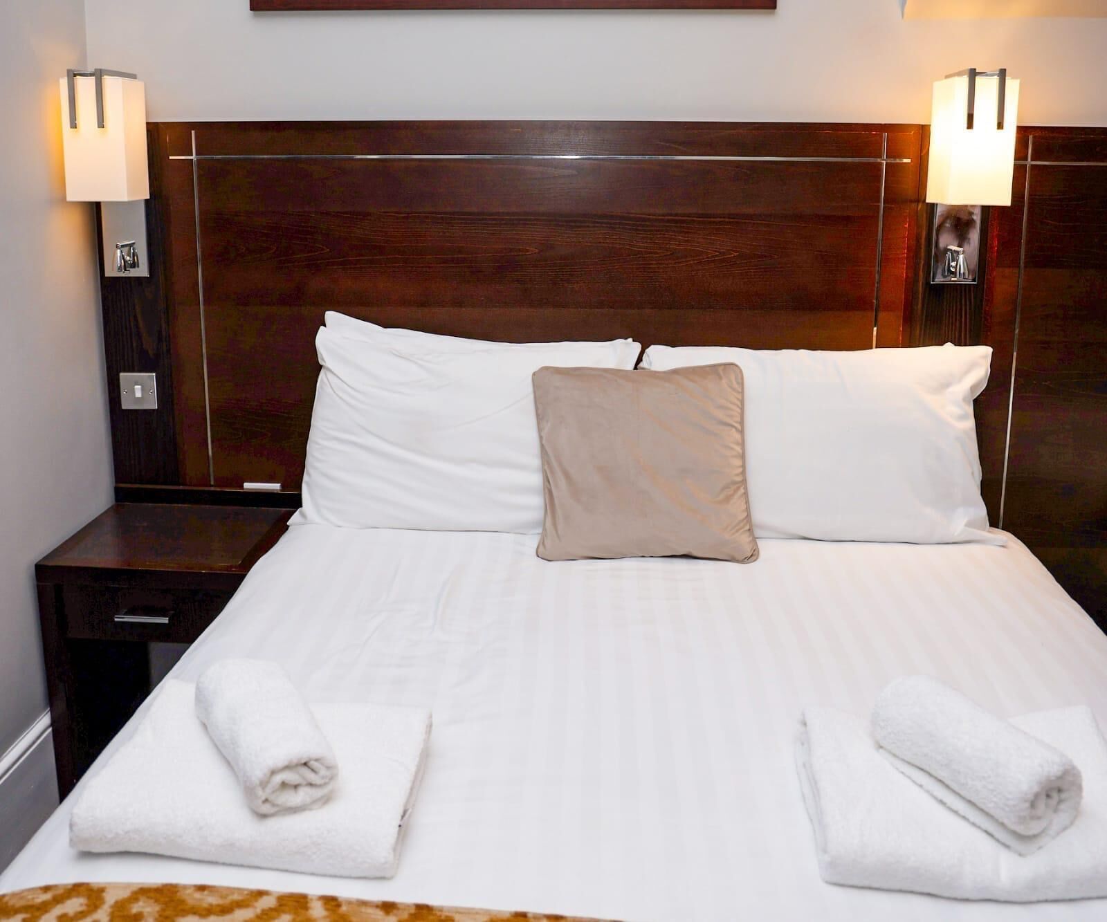 Una cama bien hecha con sábanas blancas, un cojín color beige y dos toallas blancas dobladas al pie de la cama.