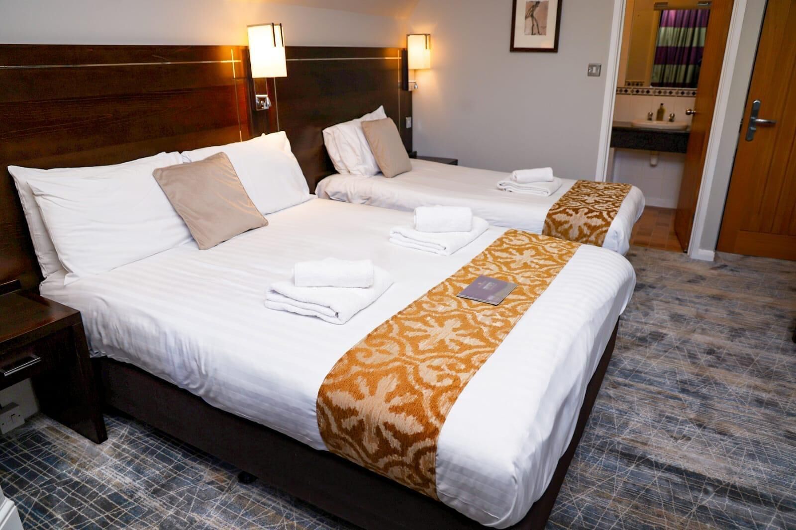 Une chambre d’hôtel avec deux lits, chacun agrémenté de draps blancs et d’un chemin de lit décoratif marron. On y trouve deux tables de chevet avec des lampes, une petite salle de bain avec un lavabo, et une porte qui donne sur une autre pièce.