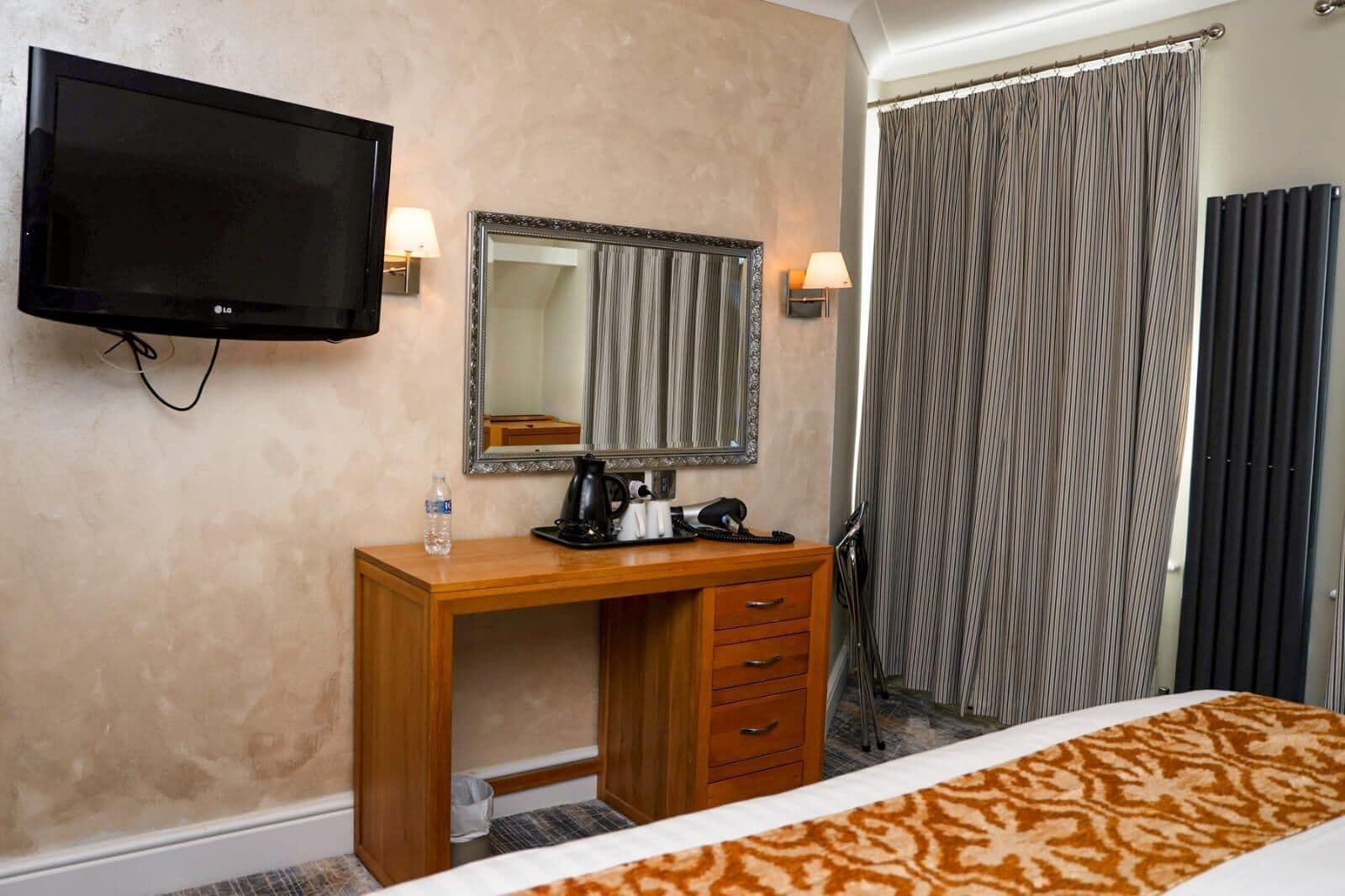 A hotel room with a bed, a wooden desk with a telephone, a mirror, a television, and a curtain.