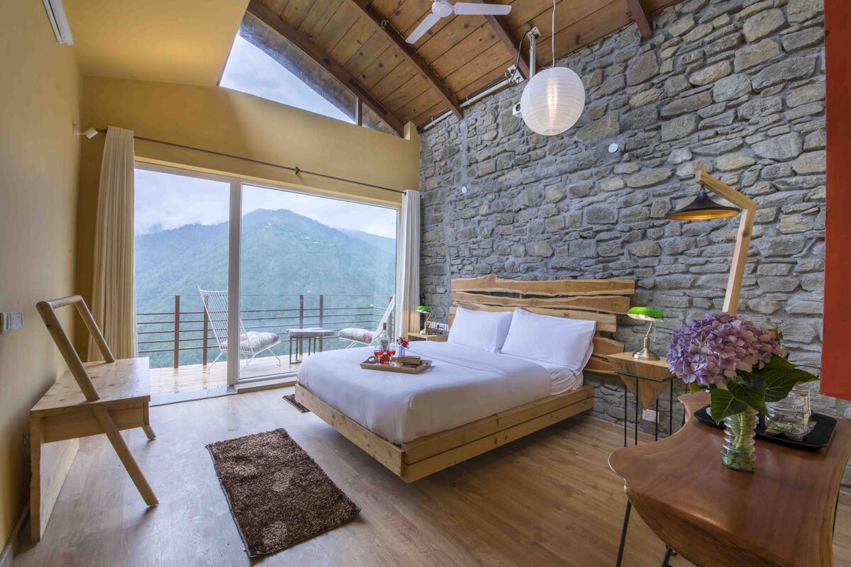 A bedroom with a wooden bed, a stone wall, a large window with a view of mountains, a wooden chair, and a nightstand with a lamp and flowers.