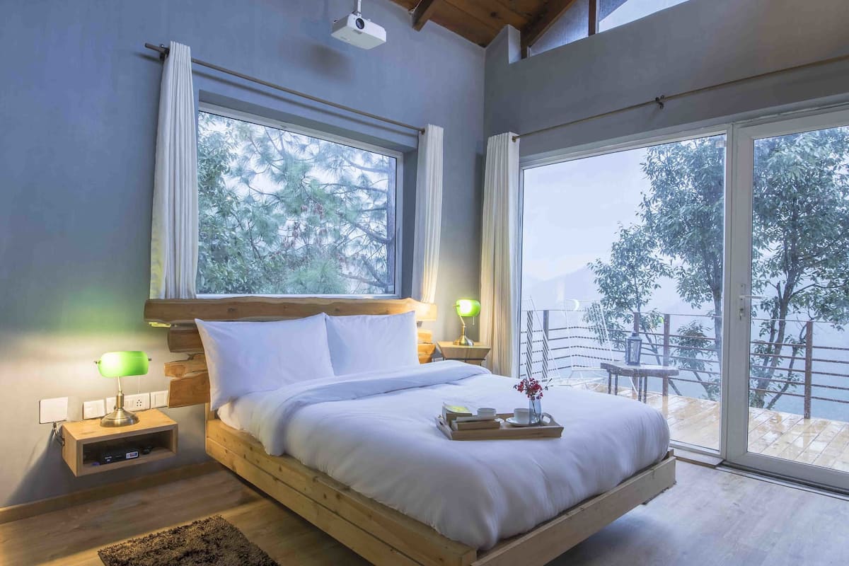 A modern bedroom with a wooden bed, large windows, and a balcony with a view of trees and mountains.