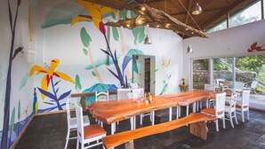 A dining area with a long wooden table, white chairs, and a mural of tropical leaves and flowers on the wall. - Seclude Ramgarh, Cliff's Edge (Kosiyakutoli)