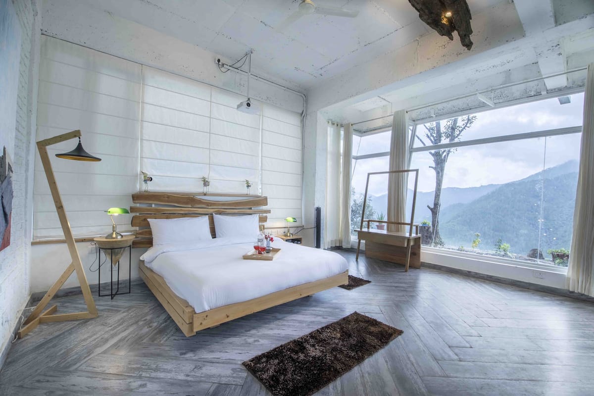 A bedroom with a large bed, wooden headboard, bedside tables with lamps, a window with a view of mountains, and a rustic ceiling.