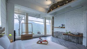 A spacious bedroom with a large bed, a wooden bench, a stone wall, and a view of mountains through large windows. - Seclude Ramgarh, Cliff's Edge (Kosiyakutoli)