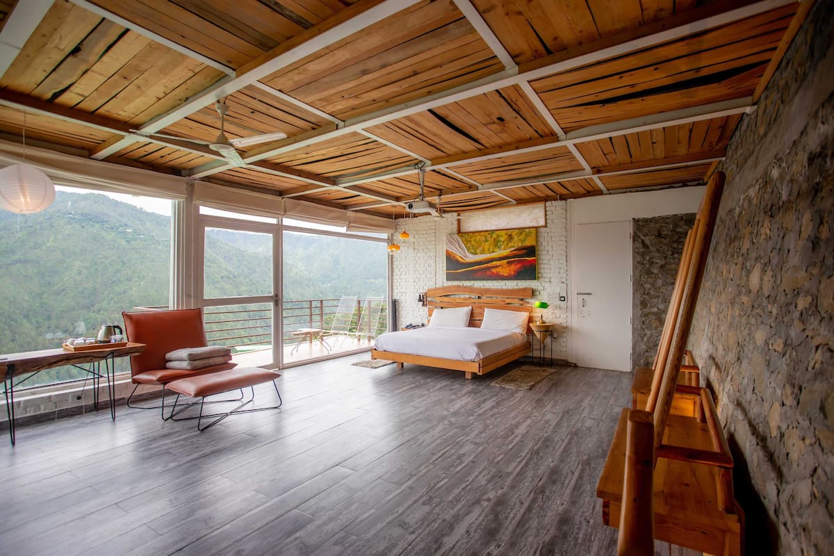 A bedroom with a wooden ceiling, a bed, a desk with a chair, and a view of the outdoors.