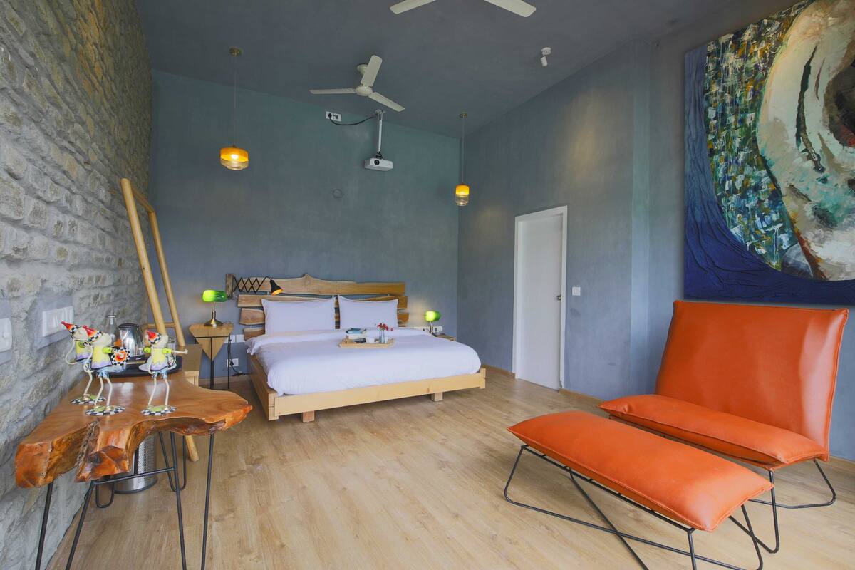 A bedroom with a bed, a wooden table with chairs, a standing fan, and a large painting on the wall.