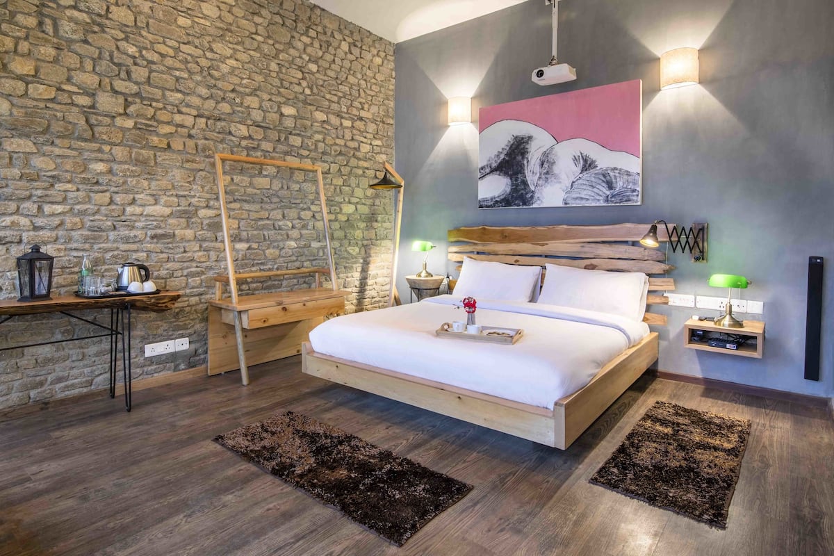 A bedroom with a stone wall, a bed with white bedding, a wooden headboard, a nightstand with a lamp, and a painting on the wall.