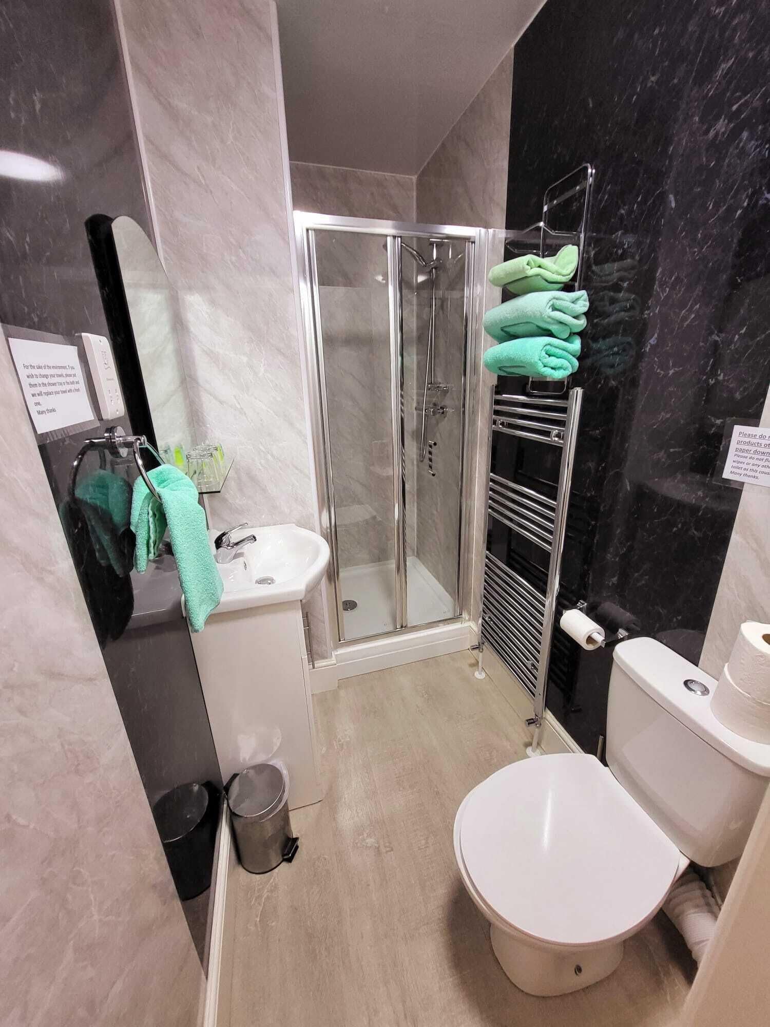 Ein modernes Badezimmer mit Duschkabine aus Glas, Waschbecken, Toilette und Handtuchhalter mit grünen Handtüchern.