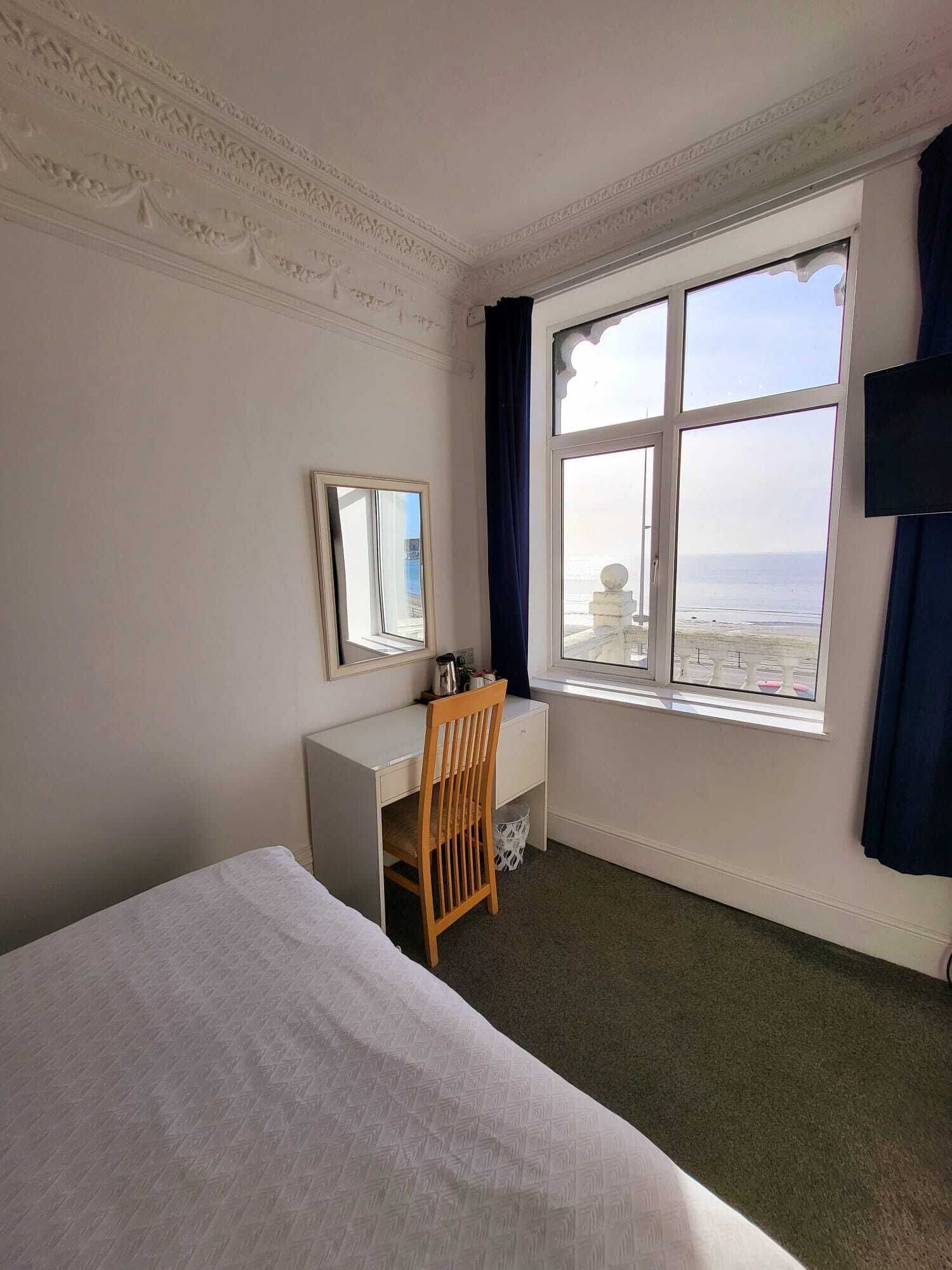 A hotel room with a bed, a desk with a chair, a window with a view of the sea, and a flat-screen TV mounted on the wall.