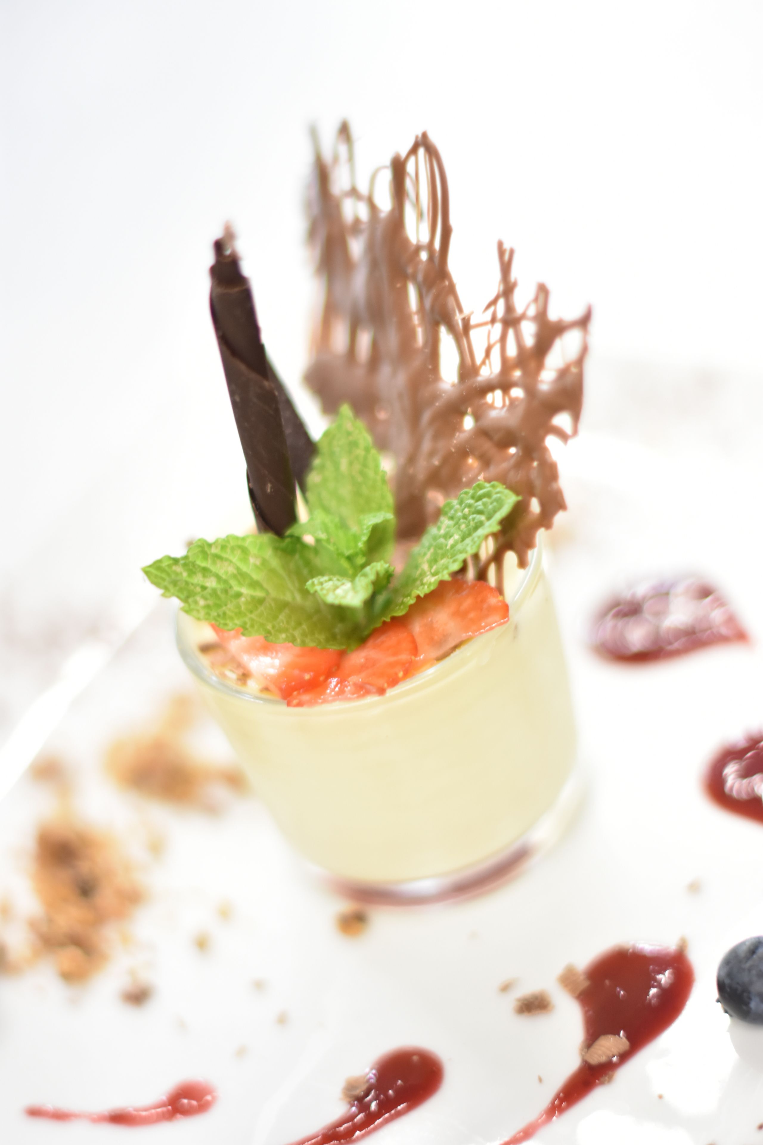 A dessert with a mint garnish, chocolate shavings, and a berry topping on a white plate with red sauce.