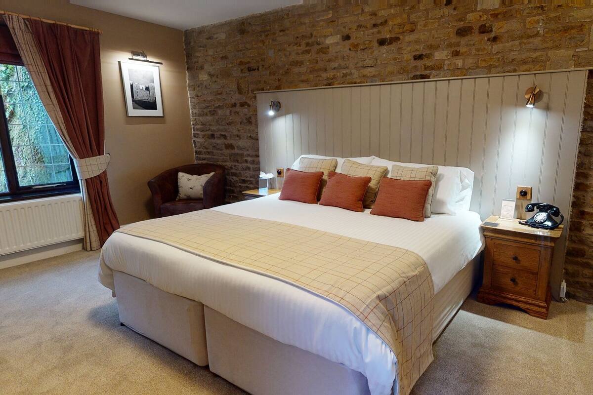 A bedroom with a large bed, a wooden nightstand, a chair, a stone wall, and a window with curtains.