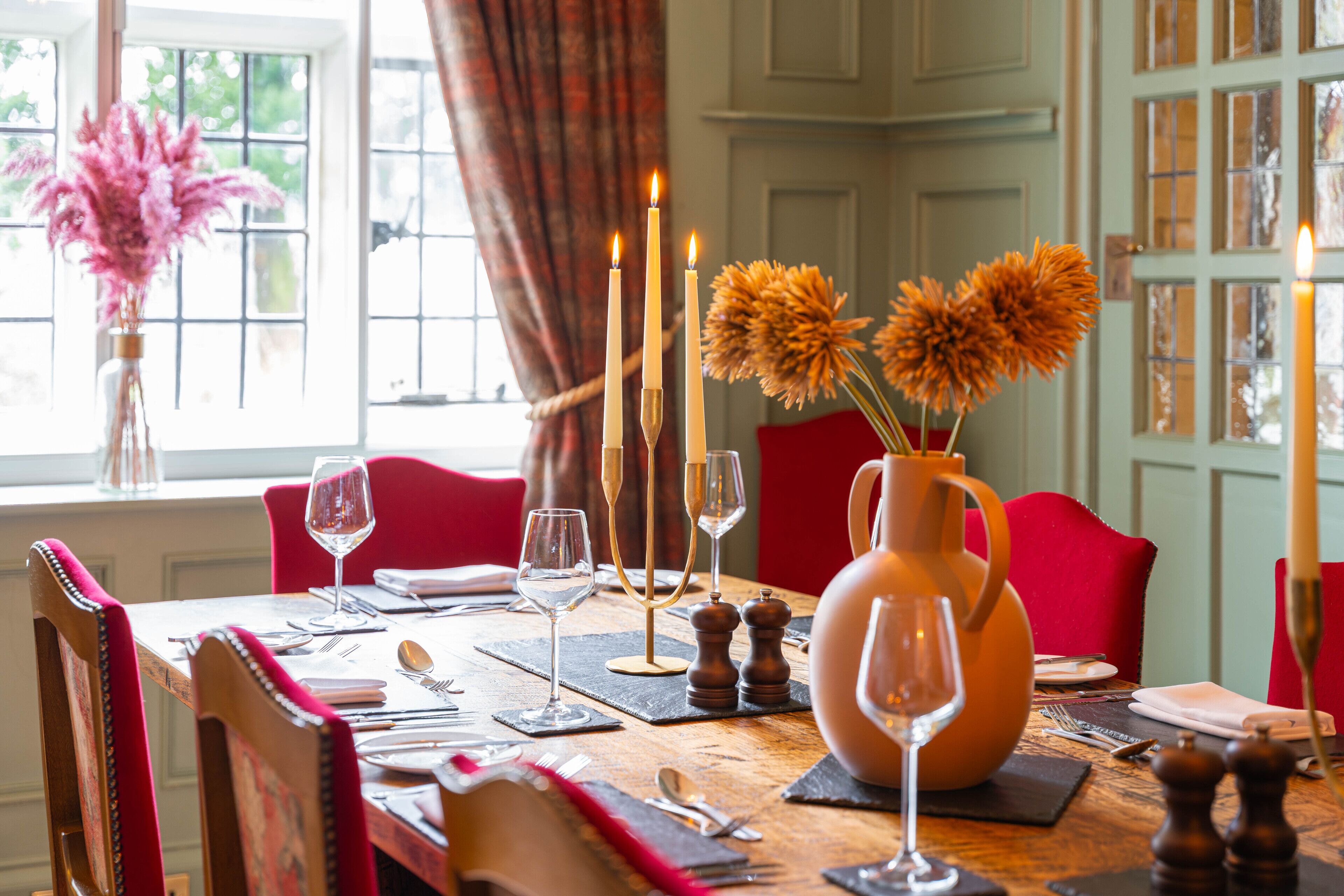 A dining table set with candles, a vase with dried flowers, and red chairs.