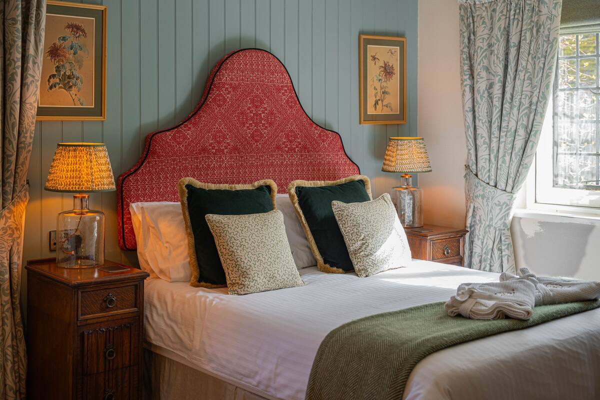 A neatly made bed with a red headboard, two bedside tables with lamps, and a window with curtains.
