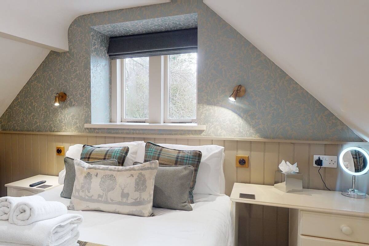 A cozy bedroom with a sloped ceiling, a window, a bedside table with a lamp, a mirror, and a neatly made bed with decorative pillows.