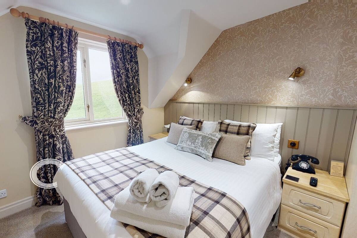 A bedroom with a bed, pillows, a bedside table with a telephone, and a window with curtains.