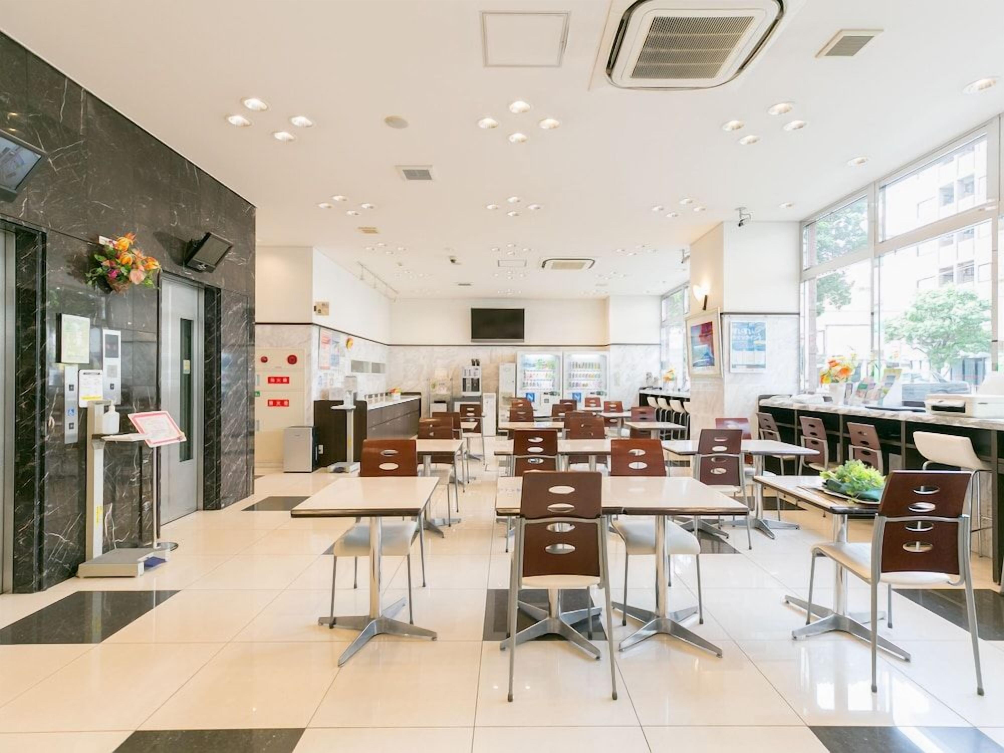 A modern restaurant with a clean, spacious layout featuring multiple dining tables and chairs, a counter area, and large windows providing natural light.