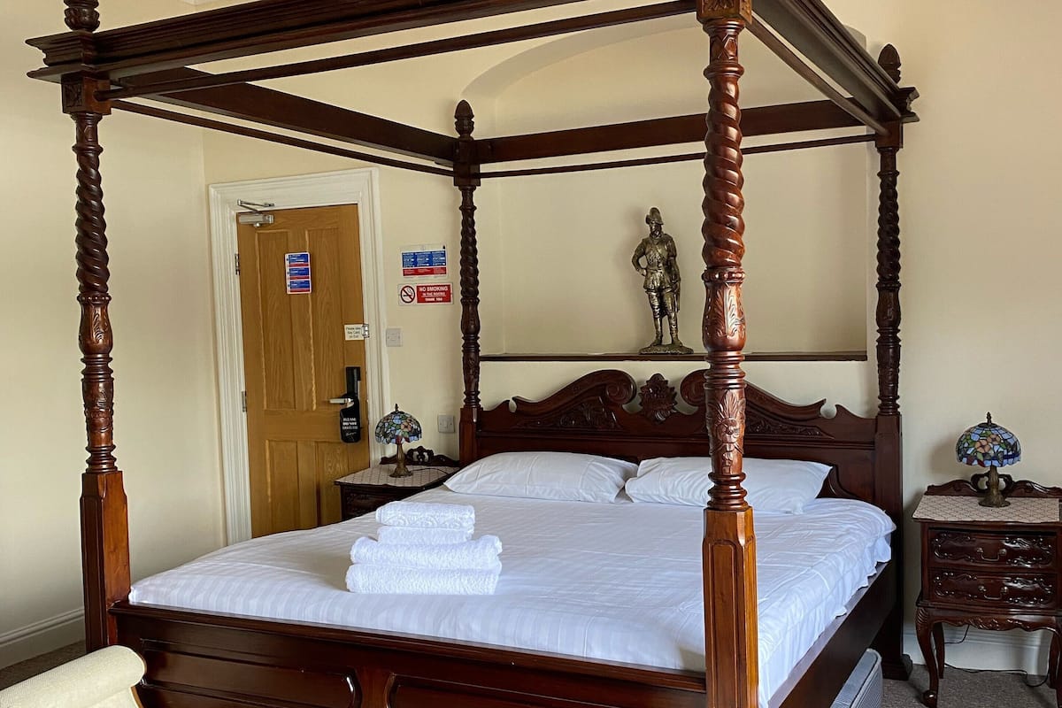 Uma cama com dossel e estrutura de madeira, um banquinho no rodapé da cama e um lustre sobre ela.