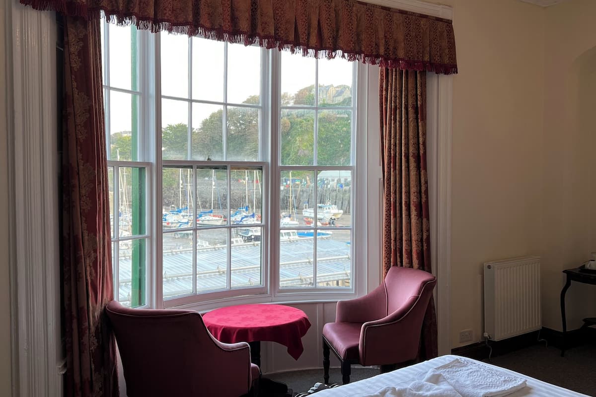Quarto de hotel com uma cama grande, duas cadeiras, uma mesa pequena e vista para barcos através de uma janela ampla.