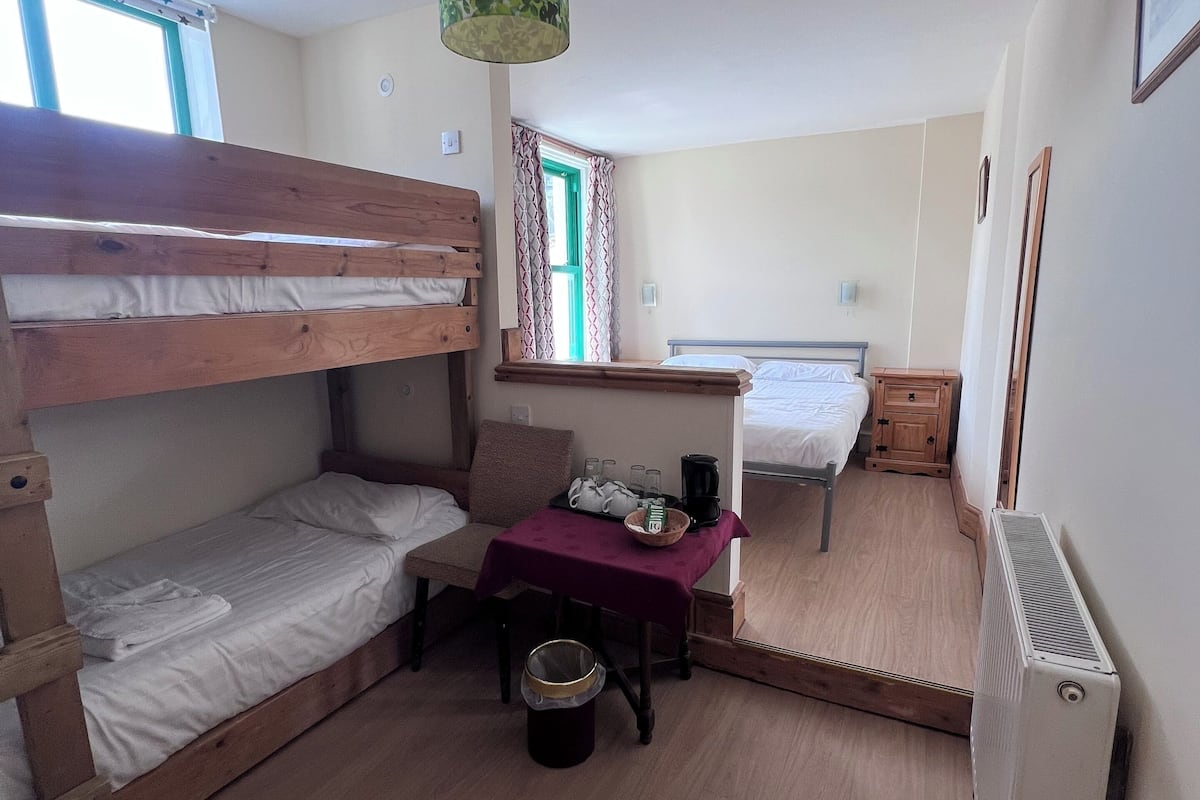 Quarto com duas beliches, uma escrivaninha com cadeira, um radiador e uma janela com cortinas.