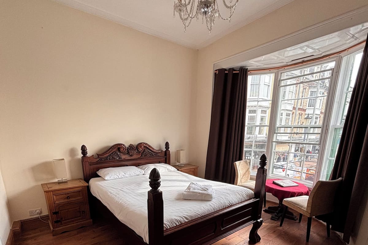 Quarto de hotel com piso de madeira, uma cama grande com cabeceira de madeira, um lustre, uma janela com cortinas e vista para a cidade.