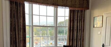 A hotel room with a large bed, a view of boats through the window, a small table, and two chairs.