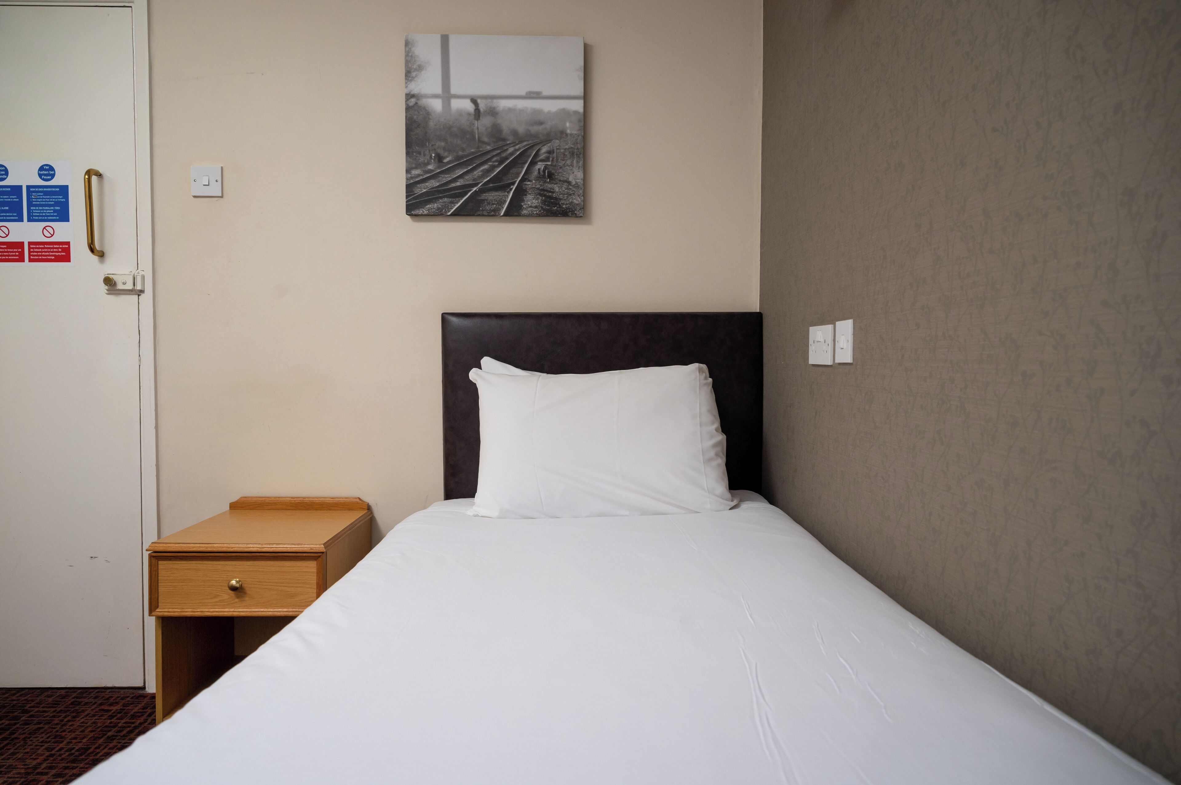 A hotel room with a single bed, a wooden nightstand, a white pillow, a wall-mounted picture of train tracks, and a door with a handle.