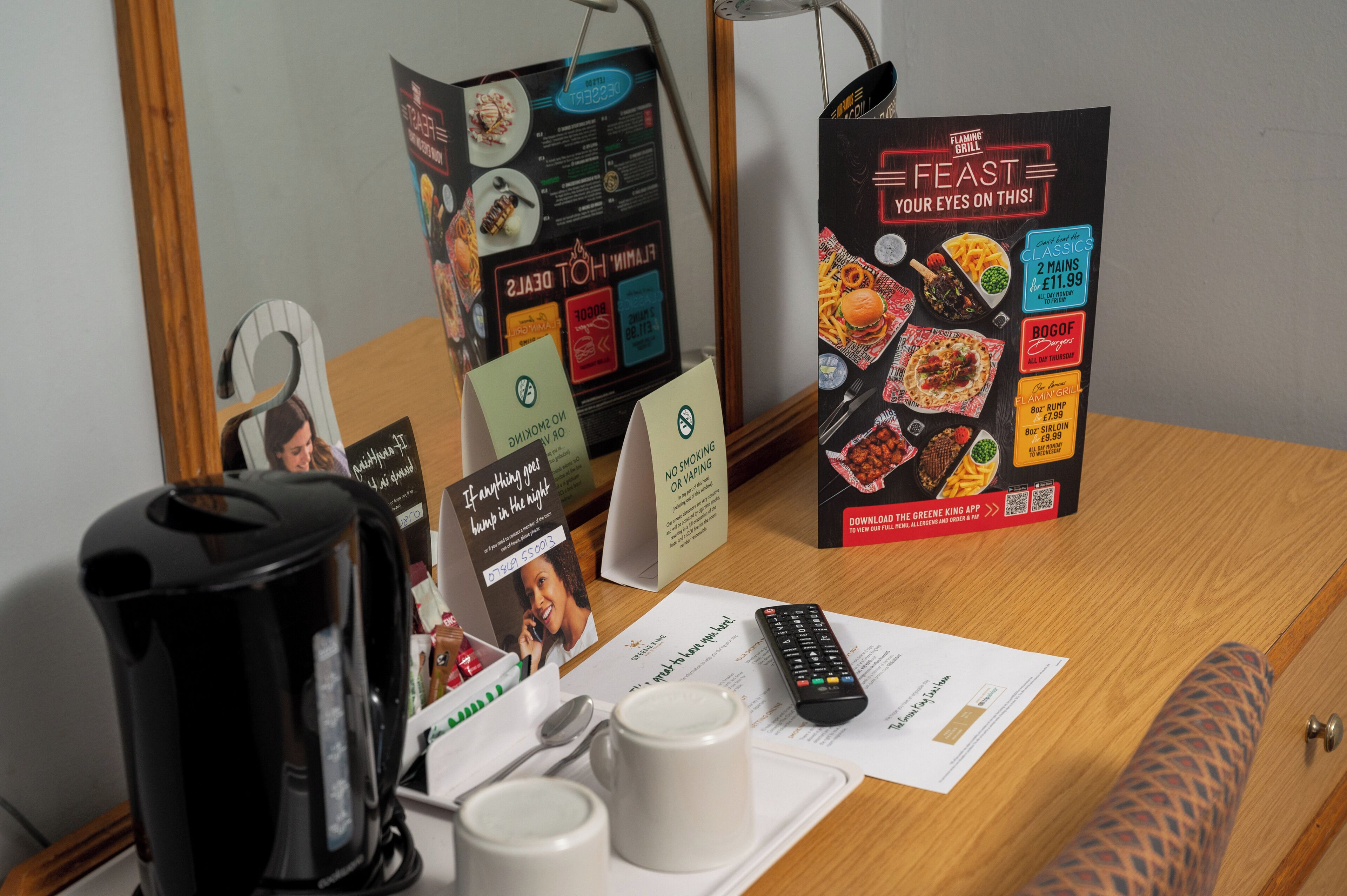 A hotel room with a wooden desk, a black coffee maker, two white mugs, a remote control, and promotional materials for the Flaming Grill.