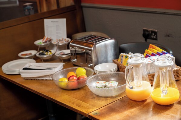 Un bufé de desayuno con diversos alimentos, un tostador y dispensadores de jugo sobre una mesa de madera.