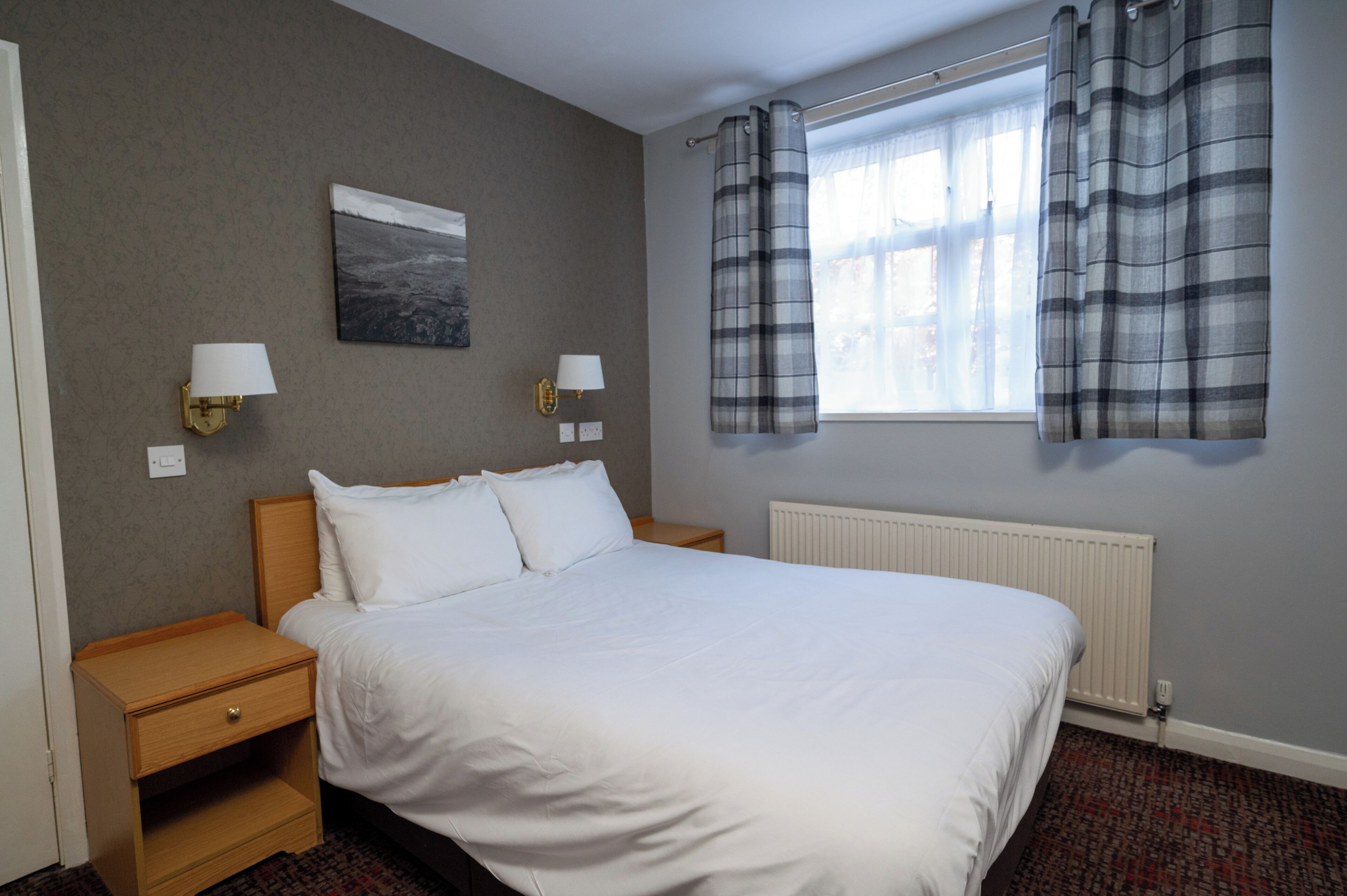 A hotel room with a large bed, two bedside tables, a nightstand, a window with curtains, a wall-mounted lamp, and a radiator.