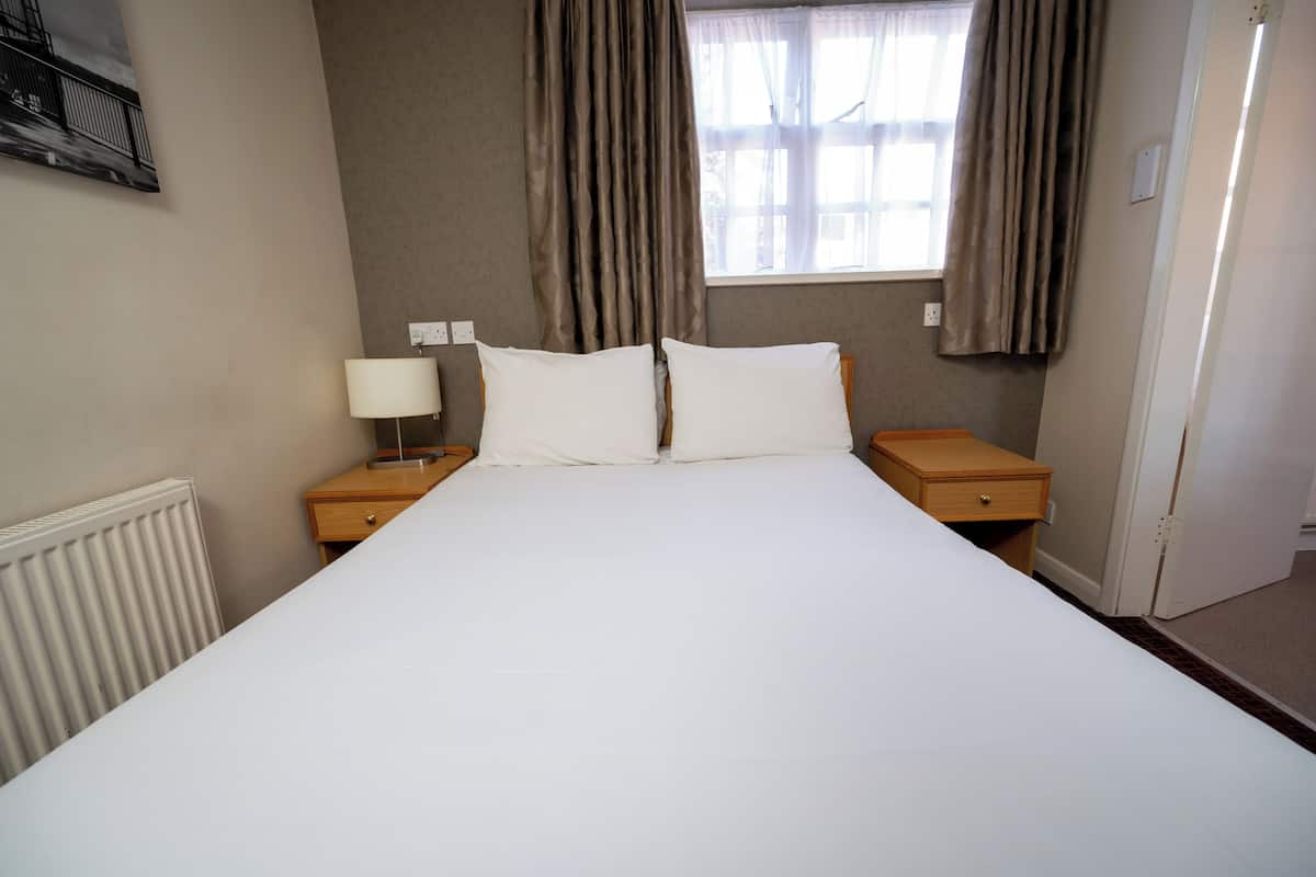 Una habitación de hotel con una cama grande, dos mesitas de noche con lámparas, una ventana con cortinas y un radiador.