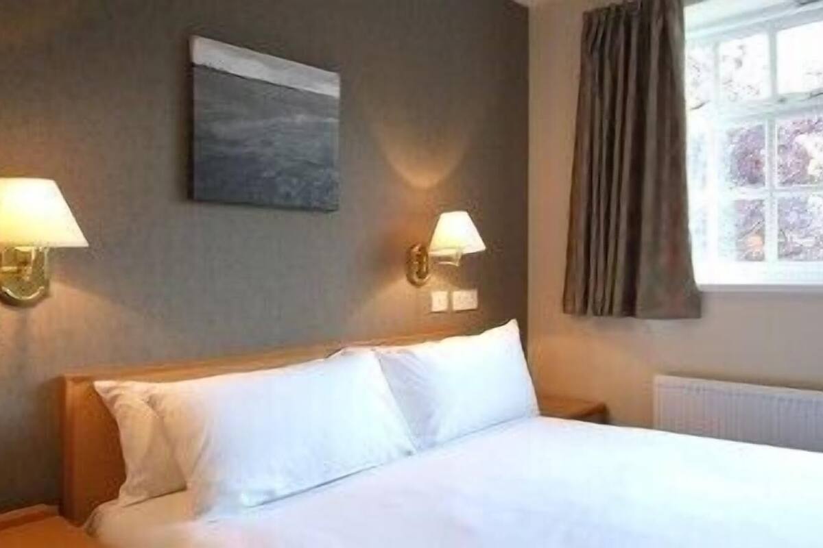 Habitación de hotel con una cama, dos lámparas de pared, un ventanal con cortinas y un cuadro en la pared.