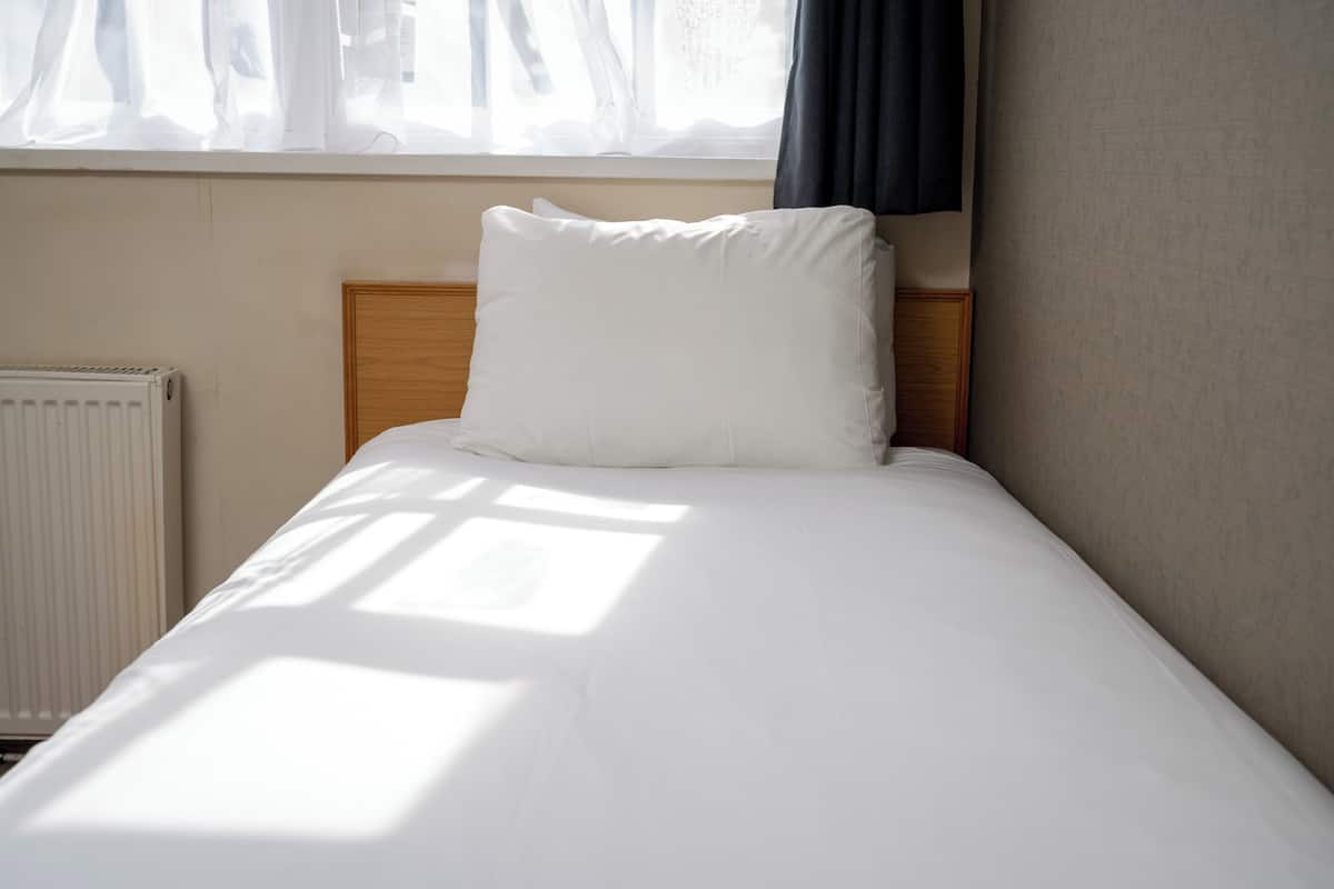 Una cama bien tendida con ropa blanca, cabecera de madera y una ventana con cortinas de gasa.