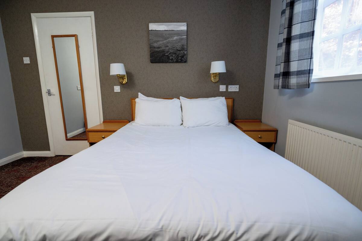 Habitación de hotel con una cama grande, dos mesitas de noche, una mesita auxiliar, un espejo, una lámpara de pared, una ventana con cortina a cuadros y un radiador.