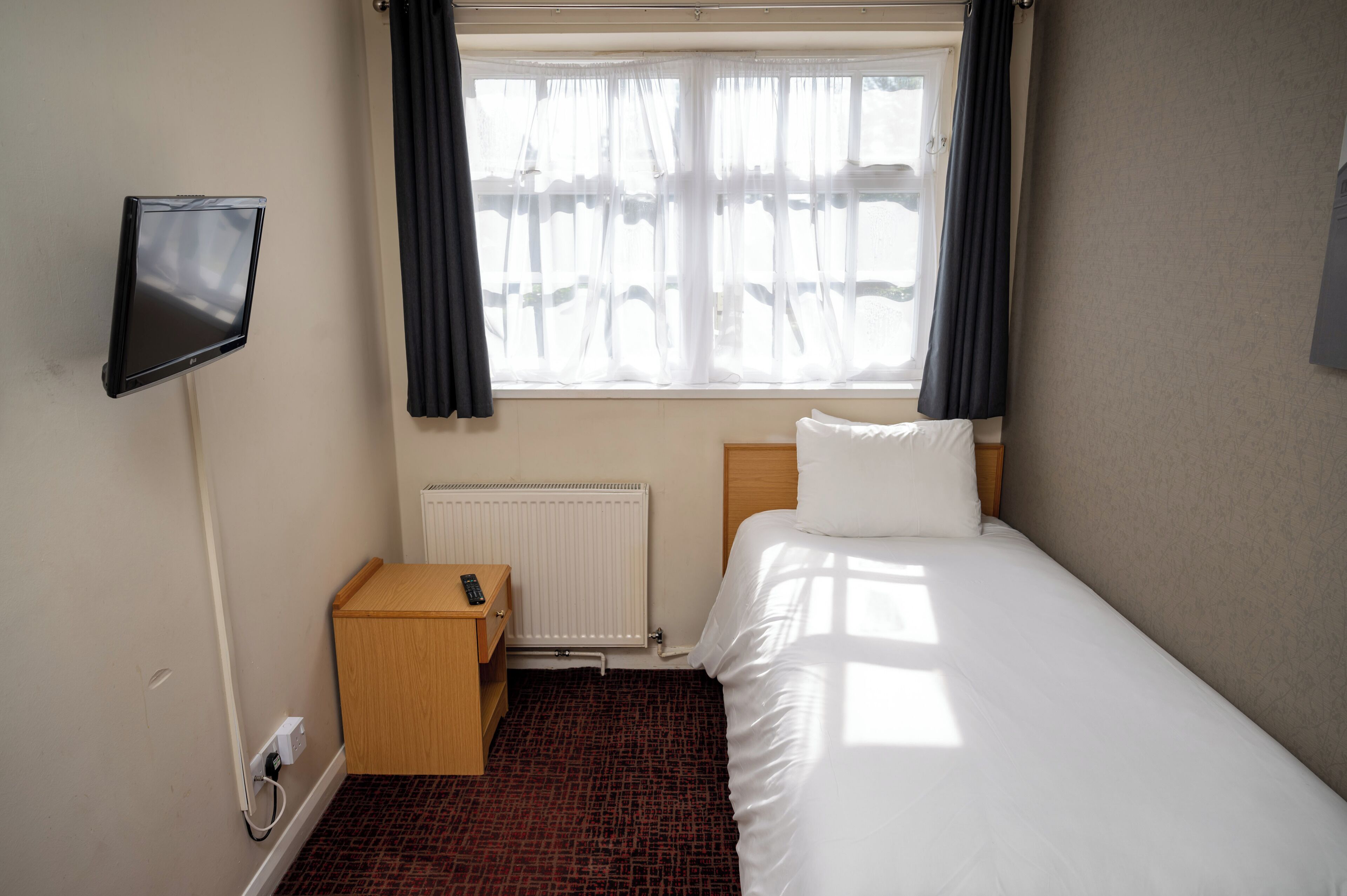 A hotel room with a bed, a TV mounted on the wall, a bedside table with a remote control, a radiator, and a window with curtains.