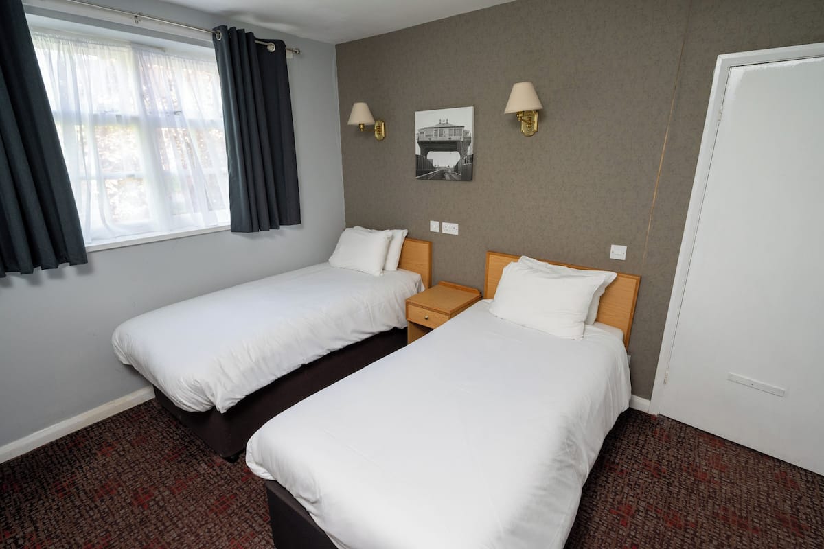 Habitación de hotel con dos camas individuales, una mesita de noche, una lámpara de pared, un cuadro enmarcado y una puerta.