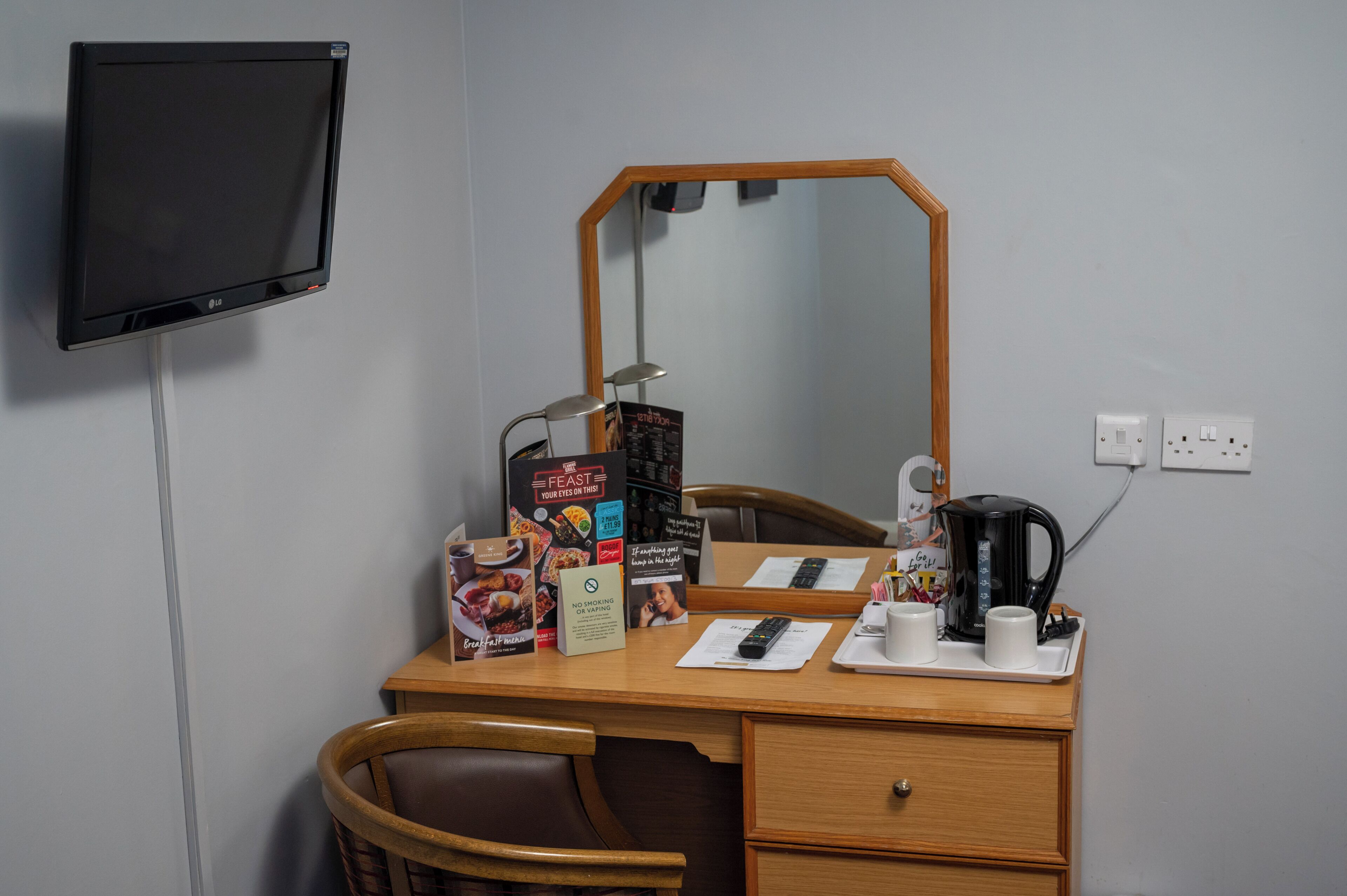 A hotel room with a desk, a television, a mirror, a coffee maker, and a tray with cups and a remote control.