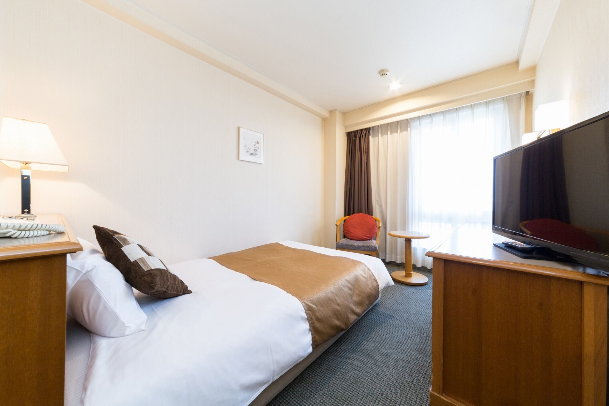 Et hotelværelse med seng, fjernsyn, skrivebord med lampe, stol og et vindue med gardiner.