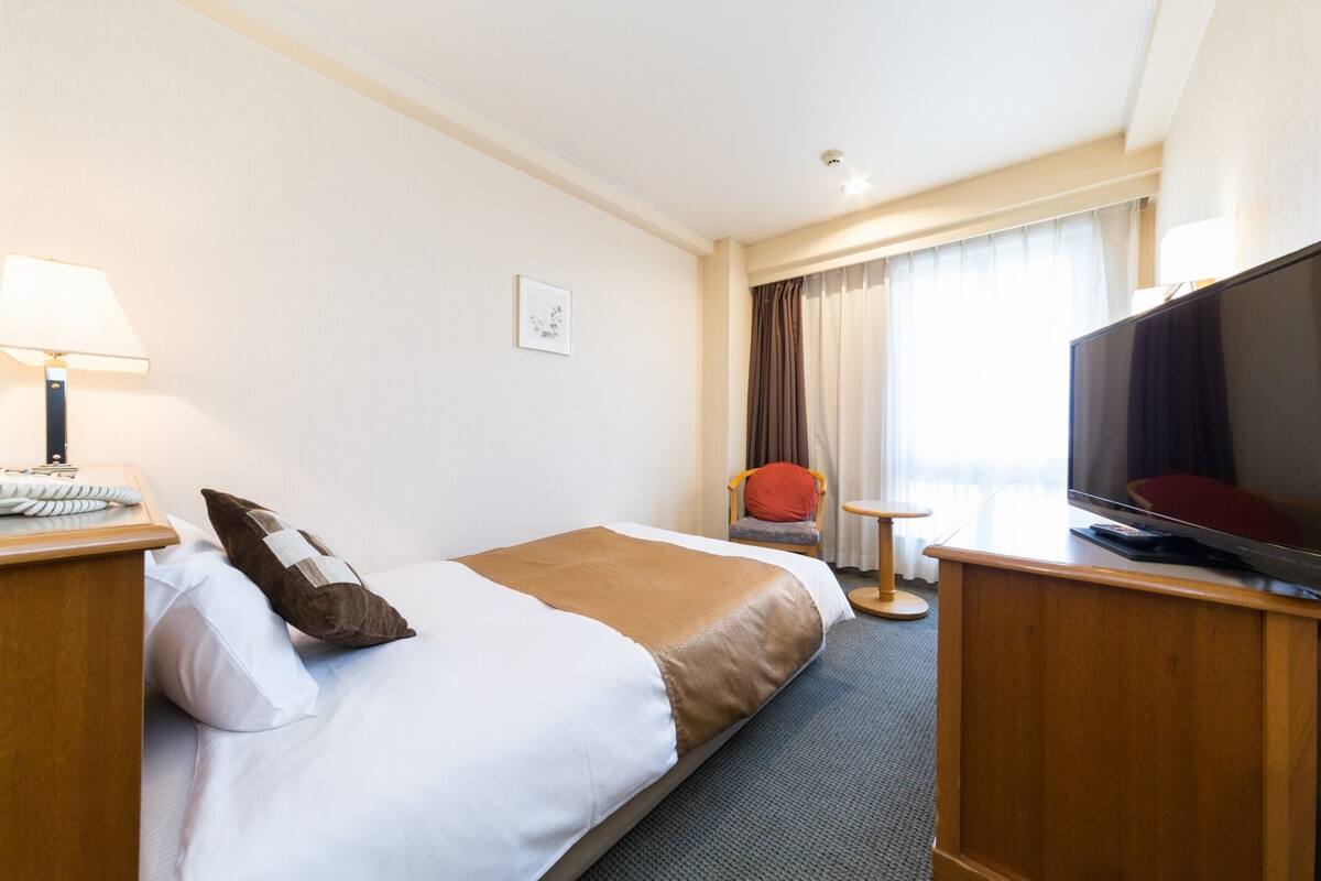 Et hotelværelse med seng, fjernsyn, skrivebord med lampe, stol og et vindue med gardiner.