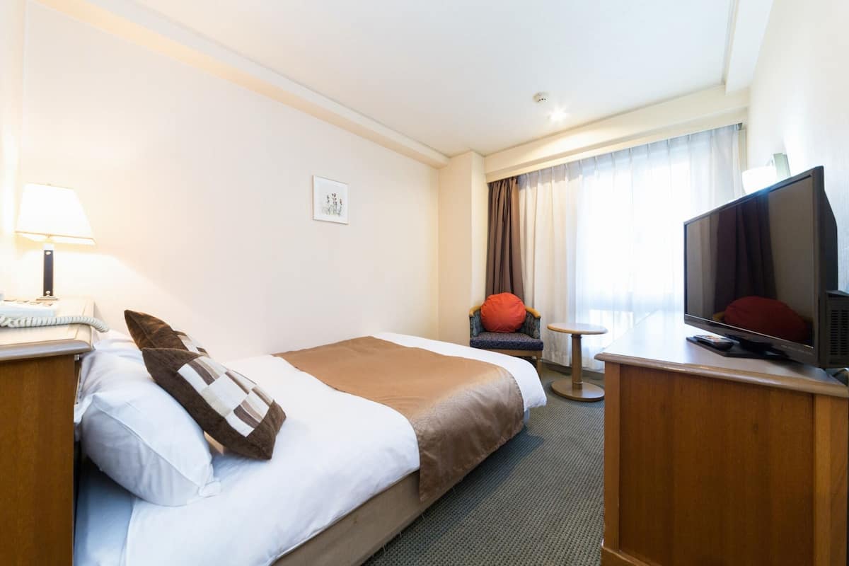 Una habitación de hotel con cama, escritorio con televisor, silla, mesa pequeña y lámpara.