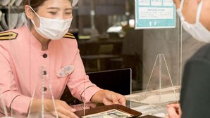 A hotel reception area with a staff member in a pink uniform behind a transparent barrier, a customer interacting with a tablet, and a sign with icons indicating hotel services. - Toyoko Inn Kawasaki Ekimae Isago (KANAGAWA)